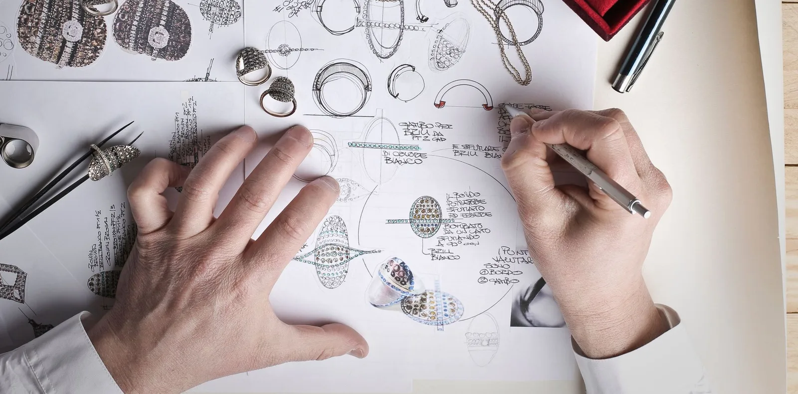 Hands drawing ring designs on sheets of sketches beside rings and tweezers