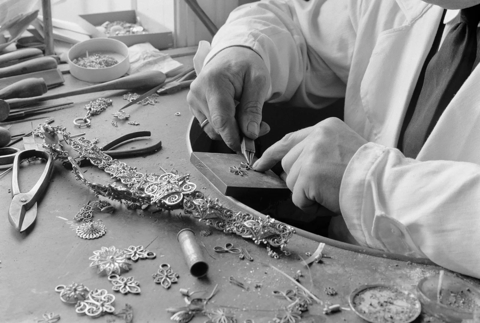 A goldsmith’s hands shaping silver filigree at a cluttered bench, in black and white