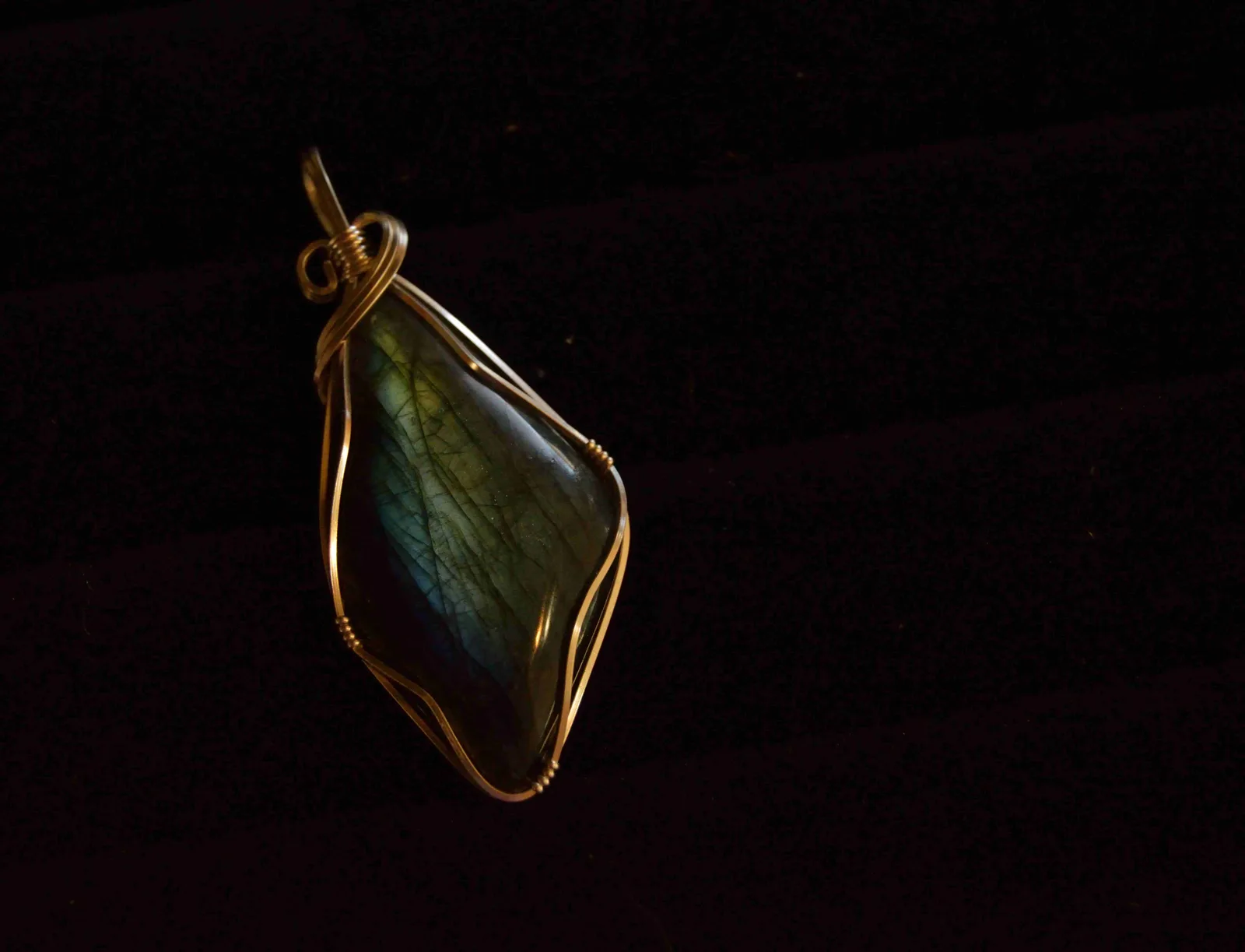 A gemstone pendant wrapped in gold wire, glowing blue and green on black velvet